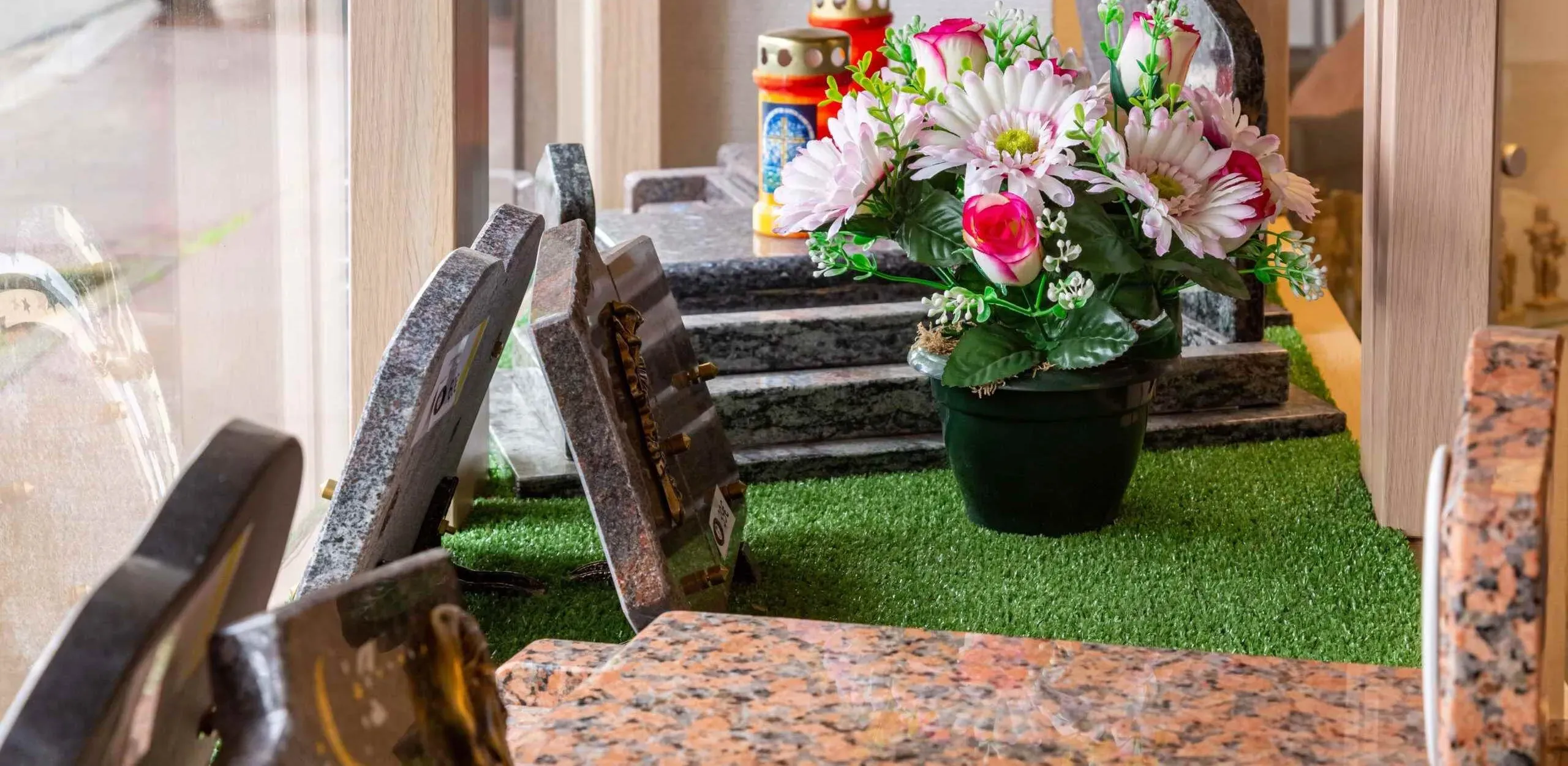 Gros plan de la vitrine avec des plaques funéraires et un pot de fleurs sur du gazon artificiel.
